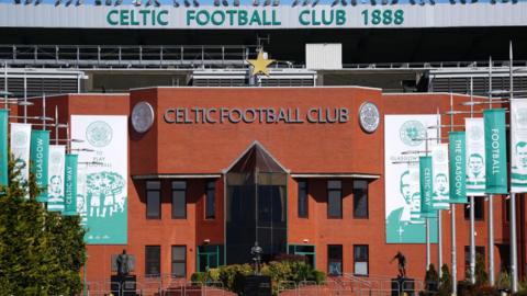 The front of Celtic's stadium. It is in red brick with green writing and the club emblem on it. There are green and white banners leading up to the entrance with former players and managers' faces on them.