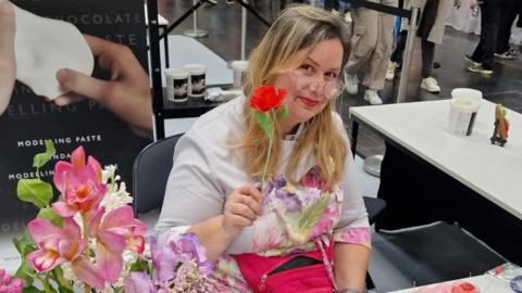 Catalina Anghel holding a rose. She is wearing glasses and has a colourful shirt on. She sits in some kind of shop behind a counter with a poster behind her with the words "modelling paste" on it.