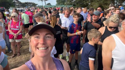 Runner Alice Harvey, who is wearing a baseball cap and a purple vest. Takes a selfie at the start line of Jersey Parkun. Hundreds or people of all ages are stood behind her. Harvey is smiling.