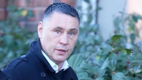 A man with short dark hair and trench coat stares into the camera.
