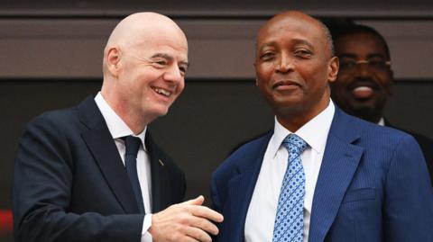 Fifa president Gianni Infantino, wearing a black jacket, white shirt and dark blue tie, smiles and holds up his right arm as he speaks to Caf president Patrice Motsepe, who is wearing a blue suit jacket, white shirt and blue patterned tie. The pair stand close together and are visible from the stomach up