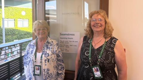 Erica Russell and Marlien Beets smile for the camera. They are stood beside an open door with the word 'seamstress' etched into its glass.