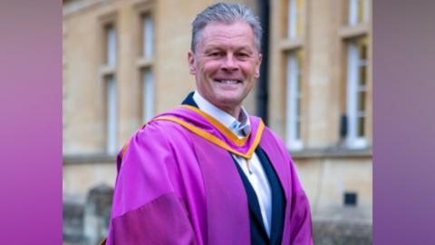Steve Cotterill, manager of Cheltenham Town FC, smiles as he stands in purple robes outside Cheltenham Town Hall. He is wearing a white shirt and suit jacket underneath.
