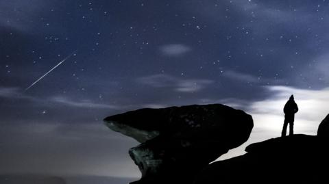 A silhouetted figure stands on rocks and watches a meteor streak across the sky