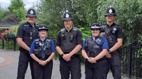 A group of five police officers, three men and two women, stand together in a pedestrianised area smiling at the camera.