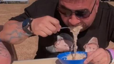 A man spoons porridge into this mouth. It is spilling into a plastic blue bowl. He is wearing sunglasses.