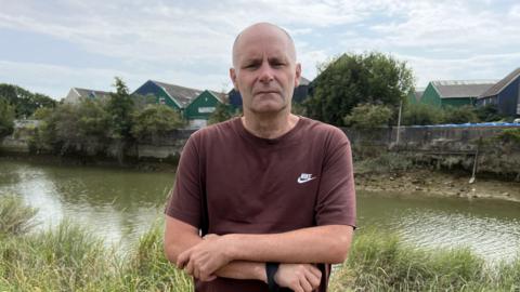 A man, Steve Gallagher, wearing a brown top. He is stood in front of a river.