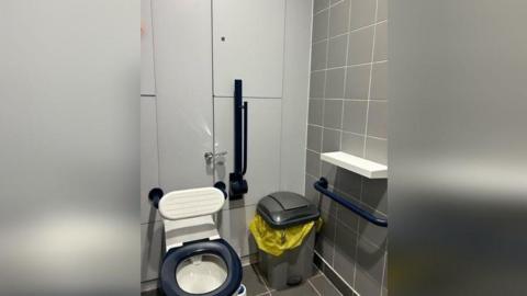 A toilet with a white backboard. A blue handrail is beside the toilet and a white shelf is attached to the wall next to it. A bin is in between the toilet and shelf.