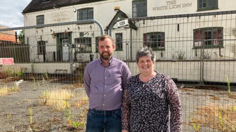 A man and a woman standing in front of a derelict pub, there is a metal fence and the car park in front is overgrown with weeds