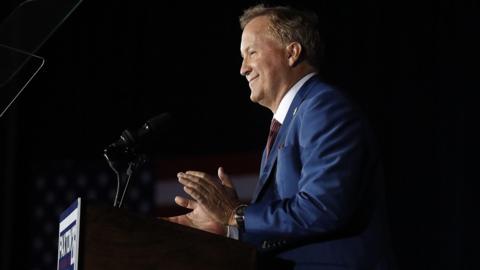 Attorney General Ken Paxton is seen in profile at a podium with a dark background from the waist up. He appears to be smiling and clapping and his campaign sign is on the front of the podium. 