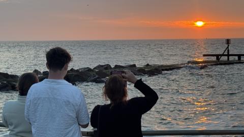 A view of a sunrise over a sea, watched by three people with phones held up filming it