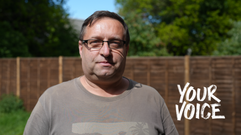 Mark Sumner looking into the camera with a neutral expression standing in a garden with a wooden panelled fence behind him with grass, trees and blue sky visible