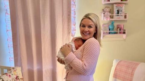 A young woman with blonde hair wears a pink knit sweater and smiles while holding a small baby on her chest. In the background is a white lounge chair with a pink blanket and the corner of a crib. 