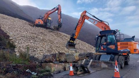 Image show mechanical diggers laying yellow stone onto a slope which had been the scene of a landslip