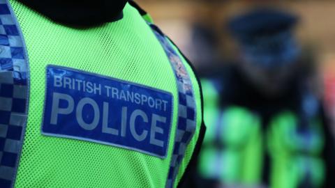 The back of a person wearing a high-visibility vest with the words “BRITISH TRANSPORT POLICE” printed on it in bold, capital letters. The vest is bright fluorescent yellow with reflective blue and white chequered strips along the sides. The text is displayed on a rectangular patch in dark blue with white lettering. In the background, there is another person also wearing a similar high-visibility vest, but this figure is out of focus, making the foreground vest the main subject of the image.