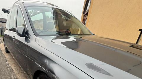 A grey Mercedes hearse parked beside a yellow wall. It has a long scratch down the side above the wheel.
