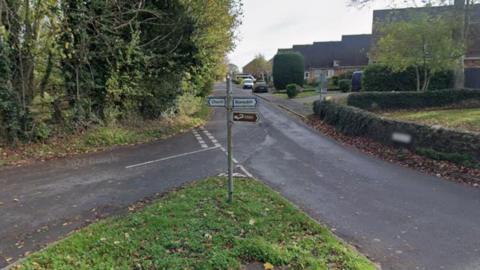 A picture taken on Blundson High Street shows street signs pointing to the right for the church and to the left for Blundson. In the background, there is a nice and quiet residential street with only a few cars parked on the pavement, and plenty of green trees and bushes.