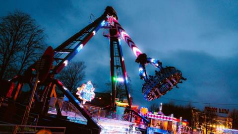 A fairground ride, lit up. Several people are strapped in and the ride is lifting them up into the air and swinging them about. 