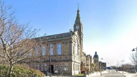 A wide photo showing the Sefton Council building. It has tall windows and a clocktower. The building can be seen lit by sunlight on a bright day.