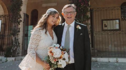 Michael Sellars, wearing a wedding suit, poses next to his daughter in her white wedding dress as she hold a large bunch of flowers