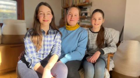 A mother sits in between her two daughters, she is wearing a blue jumper and a yellow scarf. Her daughter on the right has her hair in a ponytail and is wearing a grey jumper, the one on the left is wearing a checked shirt.