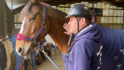 Al stands next to the horse wearing a purple hoodie and black helmet. He is stroking the horse on its neck. 