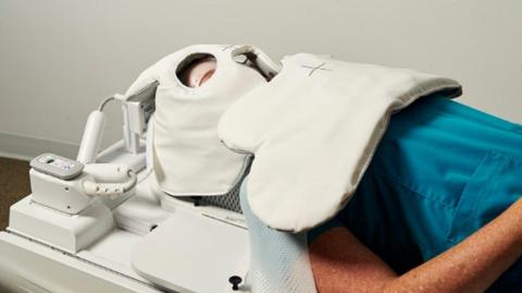 A person with their head kept in place by a white coil. They are lying on a hospital bed.