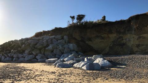 Large boulders are shown against an eroding cliff on a sand and shingle beach with bushes and the chimney of a house seen at the top of the cliff.