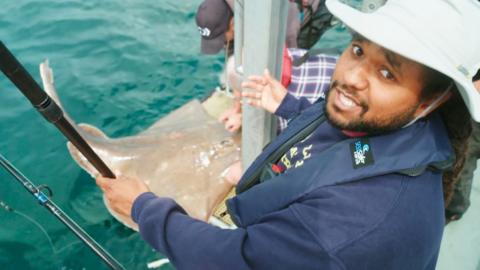 Hamza looks at the camera and notions towards a flapper skate fish in the water.