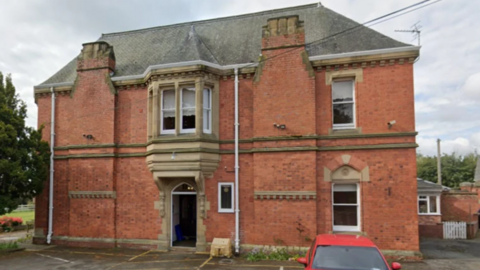 A two-storey red brick building with a sloped grey roof