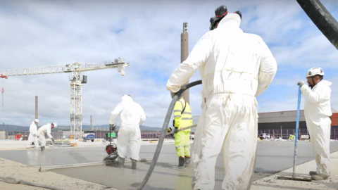 Workers wearing white suits and florescent overalls lay concrete at the treatment plant in Sellafield. There is a crane in the distance and other industrial buildings.