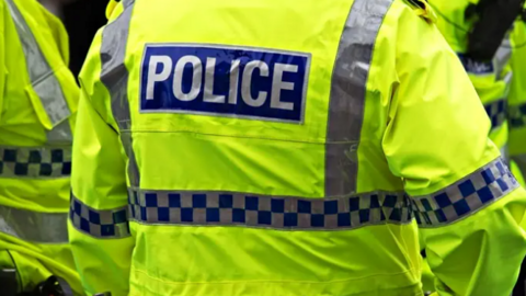 Image shows the back of a police officer's uniform. The word POLICE is in white capitals on a blue background, on a green hi-vis jacket