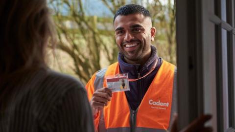 A man, smiling and  holding an ID card in his hand, at someone's door with a high-viz jacket on and fleece. There is a tree behind him. 