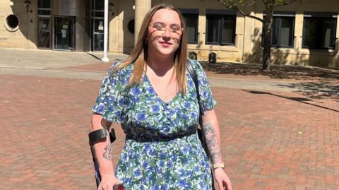 A woman in her 20s holding a crutch wearing a flowery dress outside a court building. She has tattoos on her arms, is wearing glasses and has light brown coloured hair. 