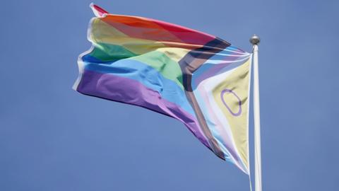 The Intersex-Inclusive Progress Pride Flag flying in the wind. It features the traditional rainbow stripes, a chevron with black, brown, pink, white, and light blue, and on the left side, a yellow triangle with a purple circle inside it.