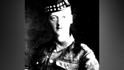 A grainy, black-and-white photo of a pictured in military fatigues. He is wearing a military-style cap. The light is striking the side of his face which appears in high contrast against a dark background.
