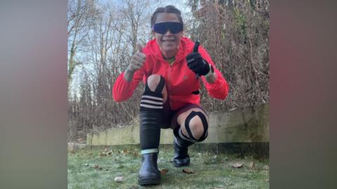 A woman crouches down in a grassy area, and gives a thumbs up to the camera. She has brown hair tied back in plaits, and wears a fluorescent pink running jacket with a hood, large sunglasses, black running shorts and black wellington boots. Her knees are strapped up with black support strips. 