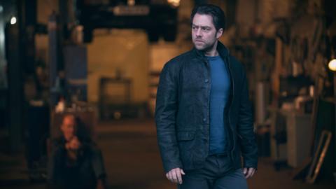 Richard Rankin, a dark-haired, brooding man in leather jacket and T-shirt looks seriously off camera. Behind him, in what appears to be a garage, a bald man is sitting on the floor, his hands apparently bound together.