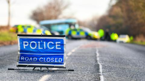 A blue sign, reading police road closed, in the middle of a road. In the background is a number of blue and yellow emergency response vehicles and police officers dressed in uniform with high visibility jackets. Behind them is  a light blue double decker bus. The background is out of focus. 