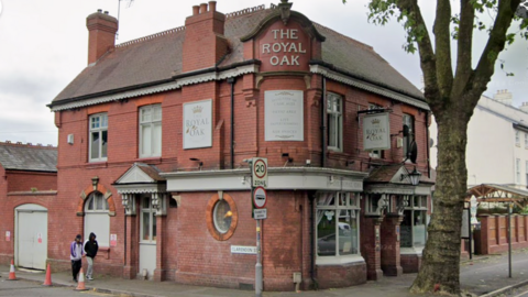 A red brick pub at the corner of two streets with a sign on the roof which reads The Royal Oak