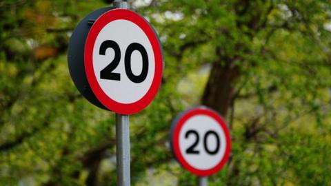 Two 20mph road signs. They are circular with a red outline and 20 in black letters.