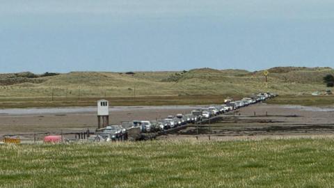 A queue of cars on a road surrounded by sand. Sand dunes can be seen in the background.