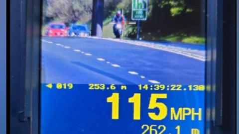 The image is of a handheld police speed‑enforcement device displaying a live camera view of a road. The screen shows a motorcyclist riding towards the camera on a multi‑lane road with a couple of cars further behind. The device readout shows a recorded speed of 115 miles per hour and a distance measurement of just over 260 metres.