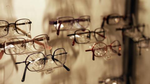 Rows of pairs of glasses lined up on display on a glass shelf at an optician's practice. Shadows cast and reflections on the glass can be seen underneath and behind them.