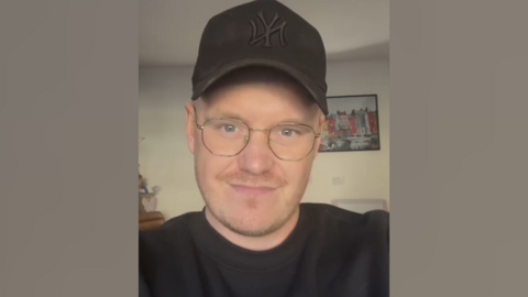 A selfie of Ryan Forey. He is wearing a black cap and top, and has rounded glasses. He is standing in front of a picture of a row of buildings. 