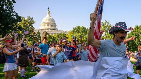 A man in a US flag hat crosses the finishing line of the Tribute to America run on the National Mall, fellow runners cheering and waving small US flags