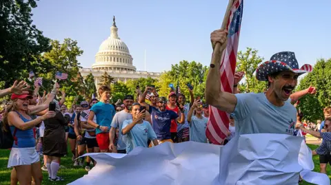 A man in a US flag hat crosses the finishing line of the Tribute to America run on the National Mall, fellow runners cheering and waving small US flags