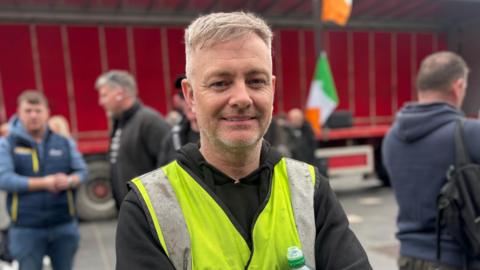 Truck driver Sonny Boyd is wearing a high-vis vest and a black hoodie. He has short hair and stubble. He is standing near a red truck with an Irish flag. Some people behind him on the street are blurred