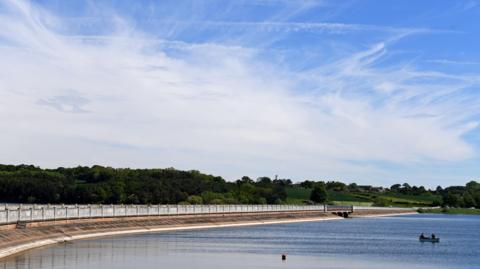 A wide view of Blithfield Reservoir in the middle of the countryside. A causeway is running along the left of the reservoir, with trees in the background. Two people can be seen in a boat on the water on the right.