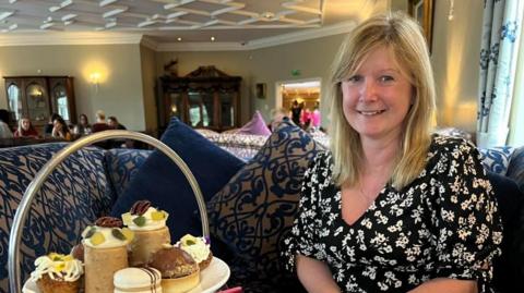 June Hoey with long blonde hair in a black and white dress with an afternoon tea tray laden with cakes beside her